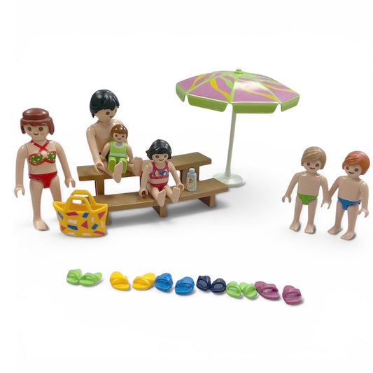 Playmobil family beach picnic set with multiple figures, a picnic table, a colorful umbrella, a yellow beach bag, and a row of colorful flip-flops on a white background.