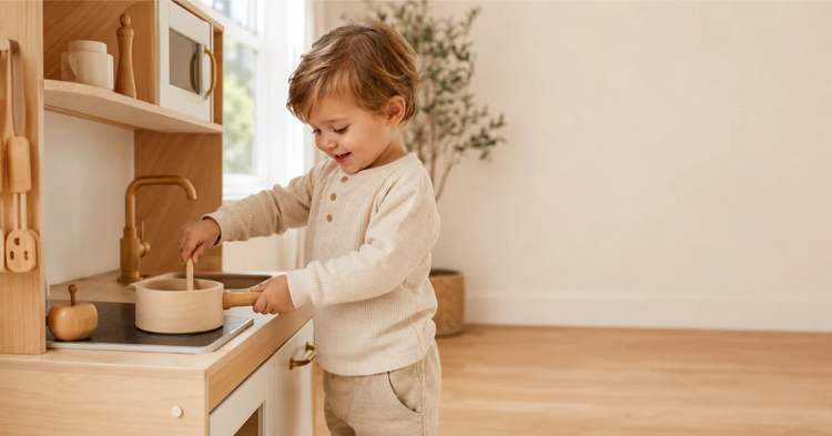 Toddler around 2.5 years standing at a wooden play kitchen, mid-action, joyful expression