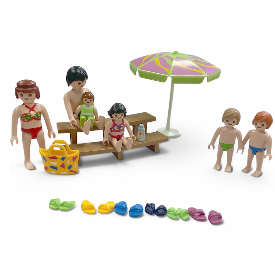 Playmobil family beach picnic set with multiple figures, a picnic table, a colorful umbrella, a yellow beach bag, and a row of colorful flip-flops on a white background.