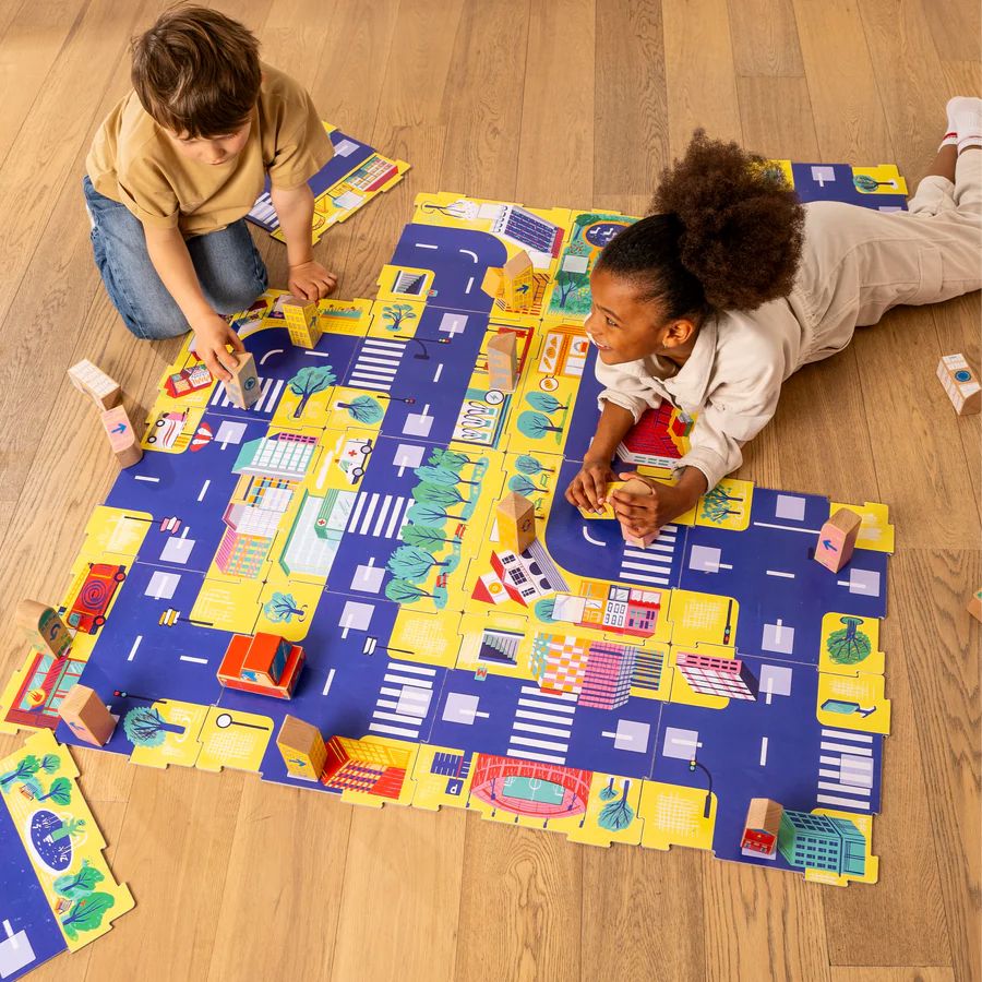 Two children playing with Cody Block City Map mat on a light wood floor. The City Map features blue roads, yellow blocks, and various illustrated buildings, cars, and trees.