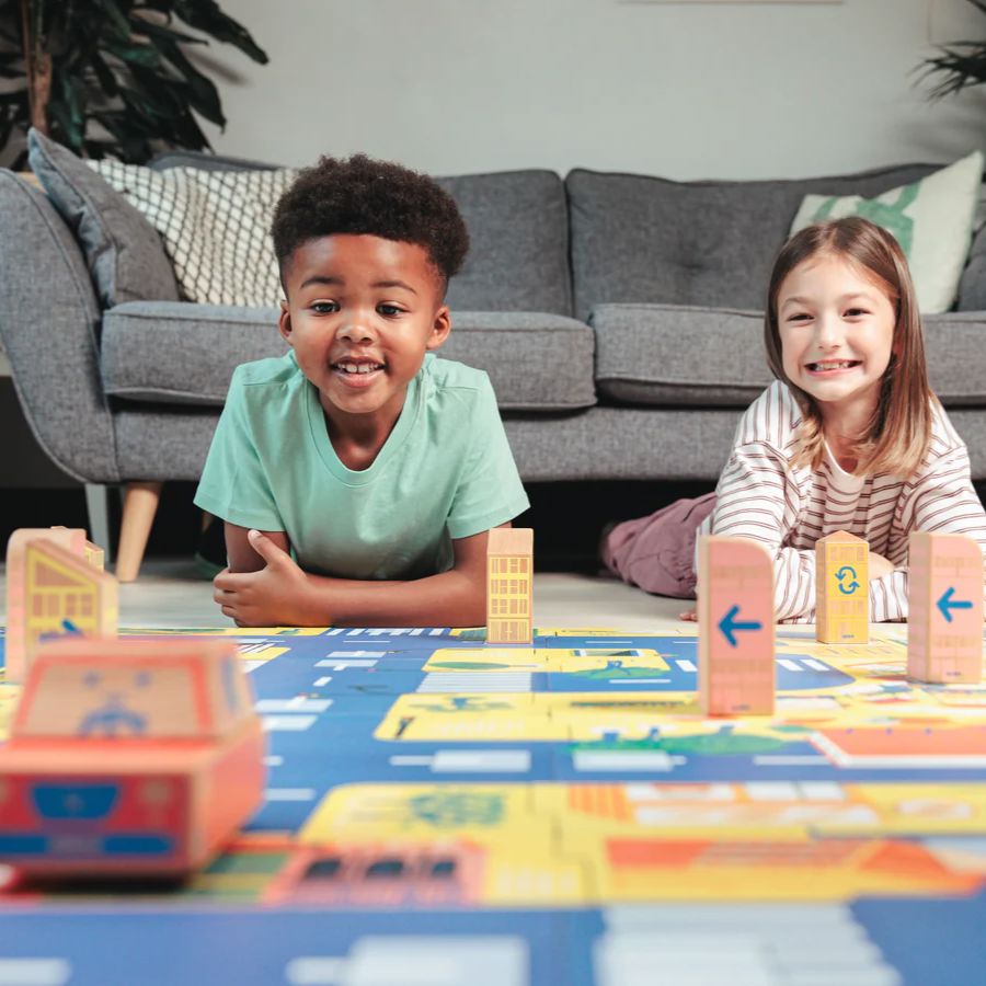 Two children smiling and playing with a Cody Block City Map and building blocks on a living room floor in front of a gray sofa.