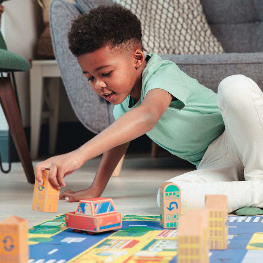 A young boy in a mint green t-shirt and white pants is lying on a blue play mat, playing with Cody Car and other cody building blocks in a living room setting.