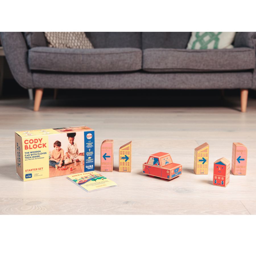 Cody Block wooden toy building set displayed on a light wood floor in front of a gray sofa. The set includes a yellow and blue box, several wooden house blocks, a red car block, and a small booklet.