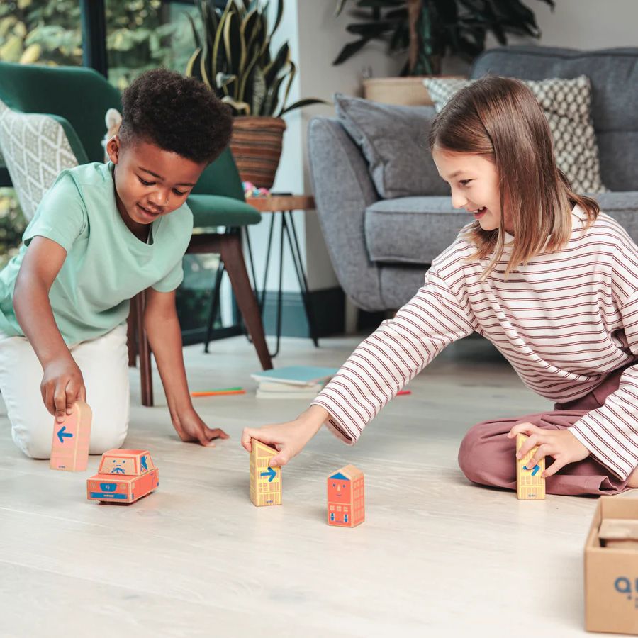 Two children sitting on a light wood floor in a living room, playing with a Cody Block wooden STEM car. 