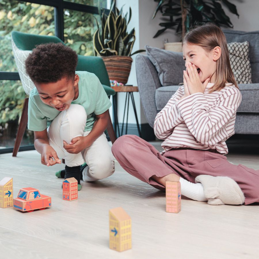 Two children playing with Cody Car and Cody Blocks  on a light wood floor in a living room. One child is crouching and smiling while building with the blocks, and the other is sitting cross-legged, covering their mouth in surprise or laughter.