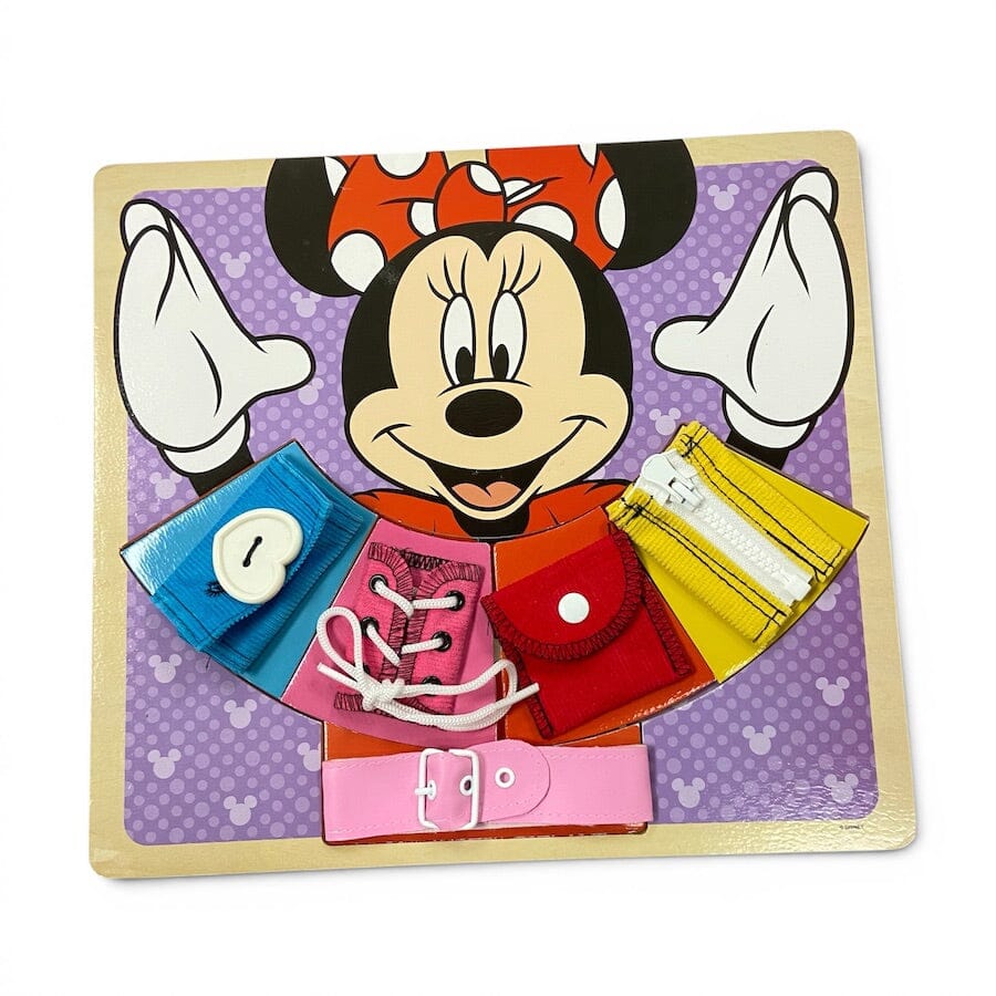 A Minnie Mouse-themed wooden toy puzzle board featuring colorful fabric accessory pieces including a blue wallet, pink shoe lace, red pouch, yellow zipper pouch, and pink belt, all arranged on a purple patterned background with a wooden frame.