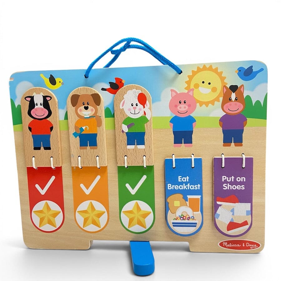 A children's wooden activity board with colorful cartoon animal figures, a blue hanging loop, and four hanging tabs showing breakfast and dressing activities. The board is photographed on a clean white background, with the Melissa & Dougno brand name visible in the bottom right corner.