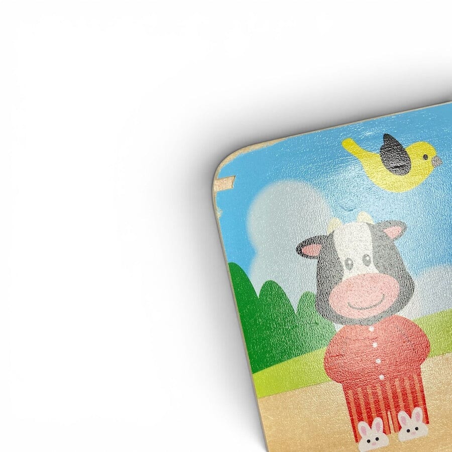 A close-up of a colorful children's toy or wooden board showing a cartoon cow in a red outfit, a yellow bird, green hills, and a blue sky on a white background.