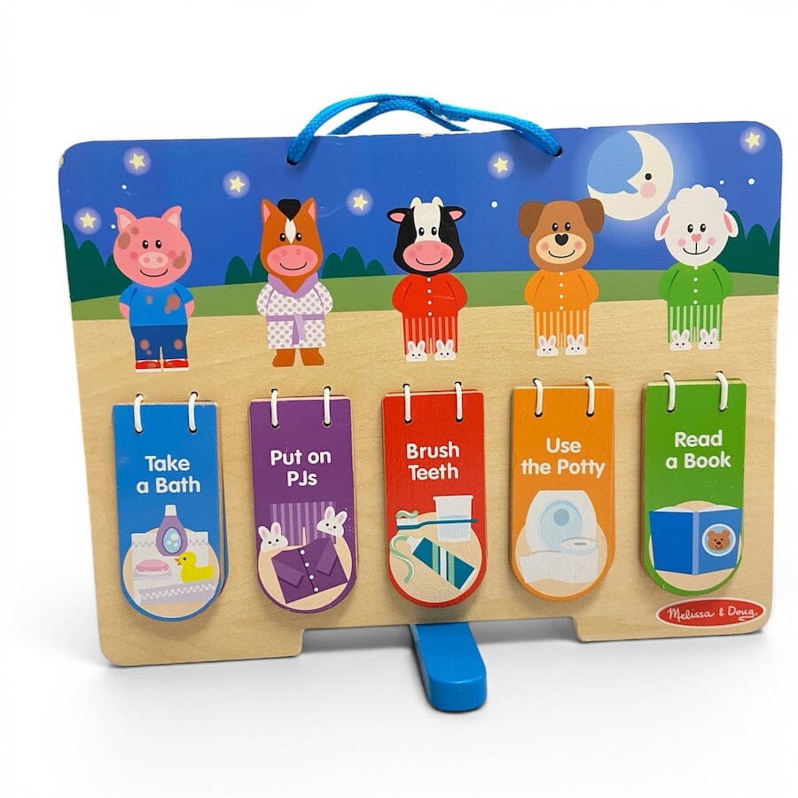 A Melissa & Doug children's wooden toy set featuring five colorful tabs with hygiene-themed illustrations and text: 'Take a Bath,' 'Put on PJs,' 'Brush Teeth,' 'Use the Potty,' and 'Read a Book.' The set is displayed on a white background with its blue carrying handle and support stand visible.