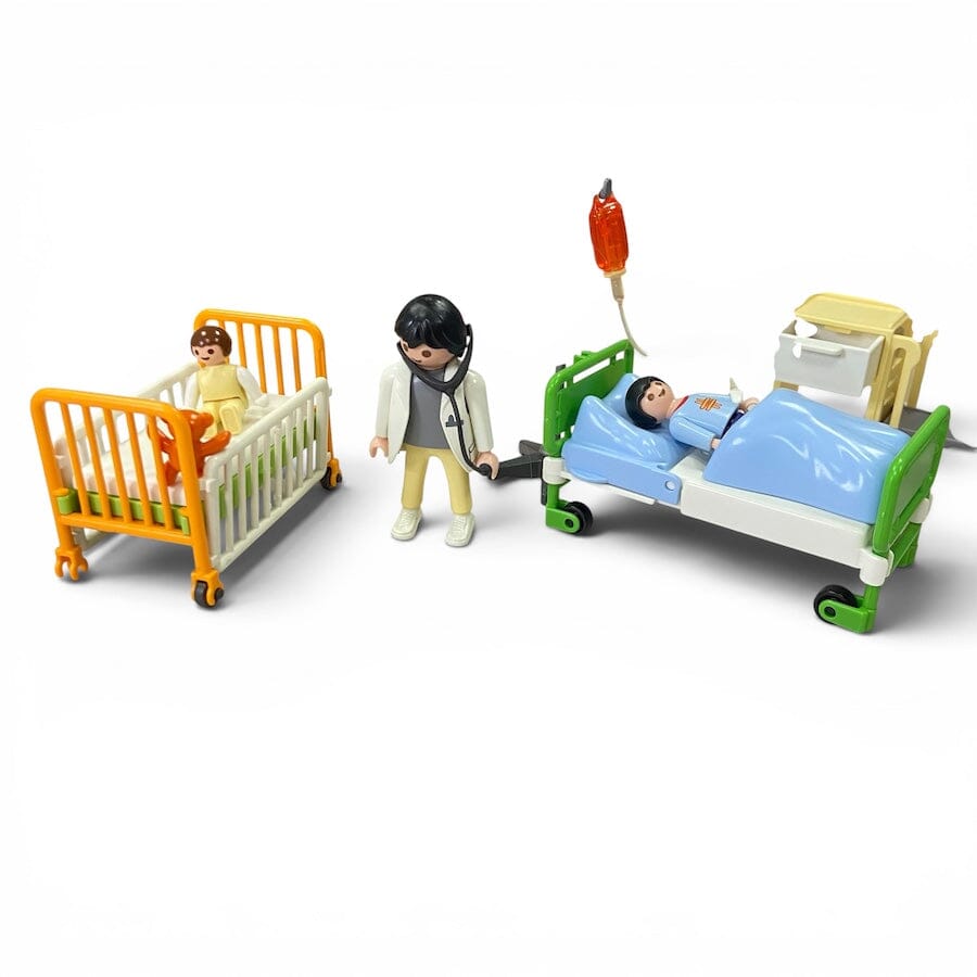 Playmobil toy medical playset with two beds, a doctor figure, and a patient figure on a white background. One bed is a yellow and white baby crib with a yellow patient, and the other is a green and white hospital bed with a blue patient under a light blue blanket.
