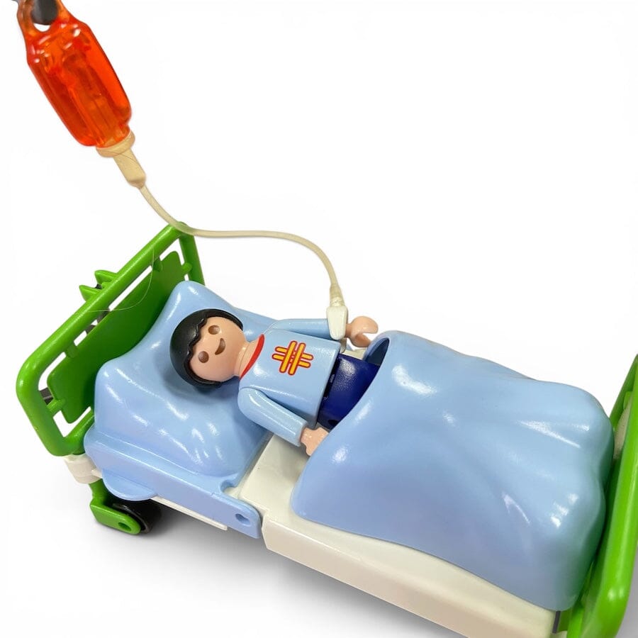 A Playmobil toy hospital bed with a patient figure lying under a light blue blanket, connected to an orange hanging IV bag on a white background.