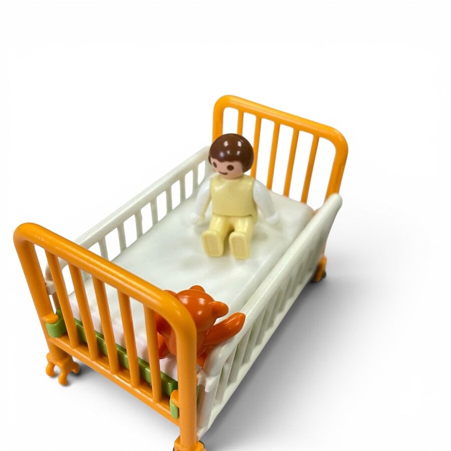 A Playmobil small toy crib with orange rails and a white mattress, shown on a white background. A seated doll in pale yellow clothing sits in the crib, and an orange stuffed animal rests near the foot of the crib.