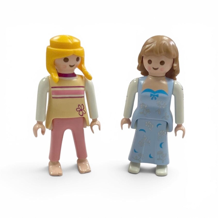 Two small Playmobil toy children's figures standing side by side on a plain white background. One figure has yellow hair and a tan top with pink pants.