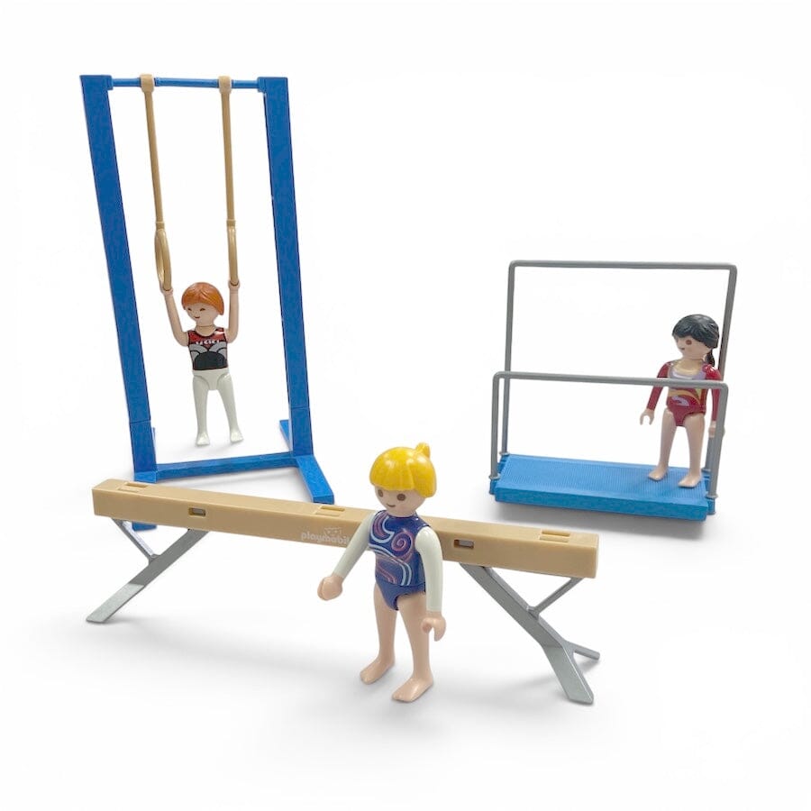Playmobil gymnastics set with miniature figures and equipment, including a balance beam, parallel bars, uneven bars, and a vaulting horse, shown on a white background.