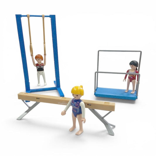 Playmobil gymnastics set with miniature figures and equipment, including a balance beam, parallel bars, uneven bars, and a vaulting horse, shown on a white background.