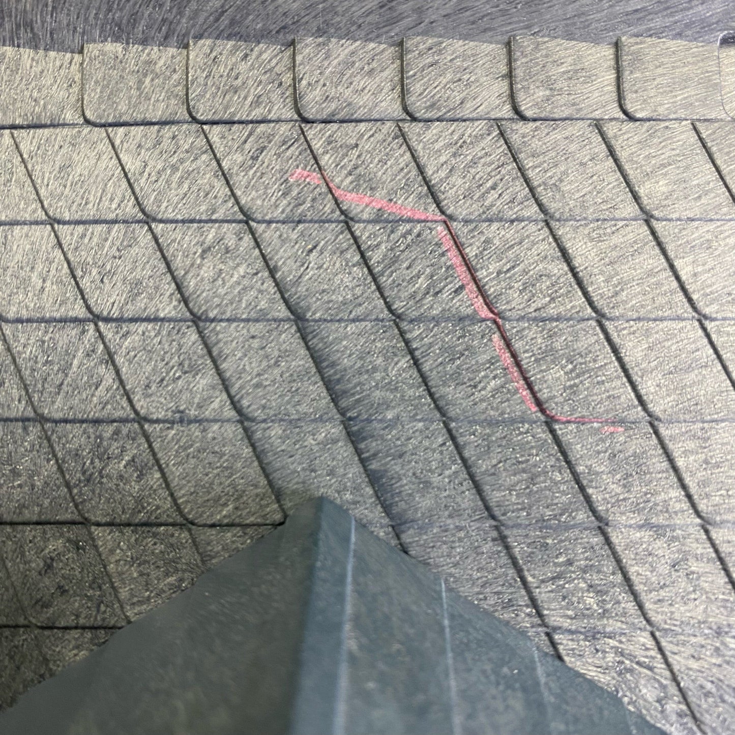 Close-up of textured gray interlocking paving stones or tiles with a red measurement marker drawn on the surface, and a dark gray corner of a board or object in the foreground.
