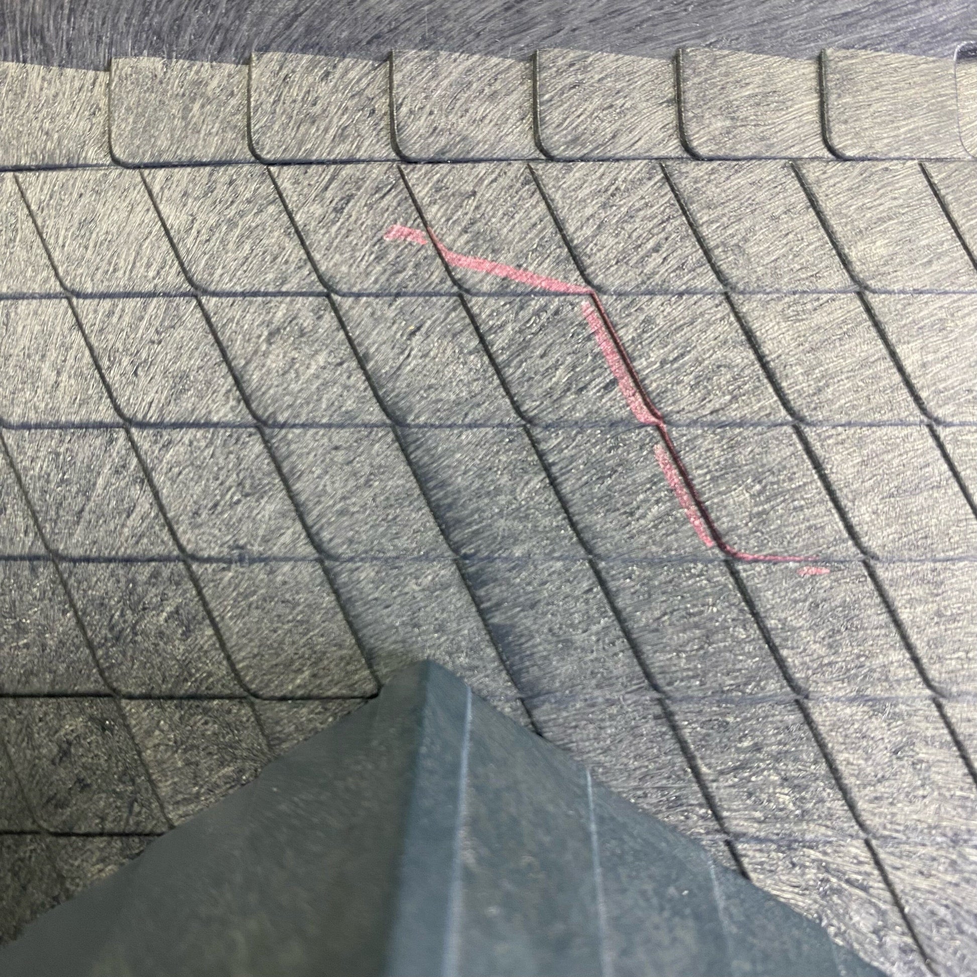 Close-up of textured gray interlocking paving stones or tiles with a red measurement marker drawn on the surface, and a dark gray corner of a board or object in the foreground.
