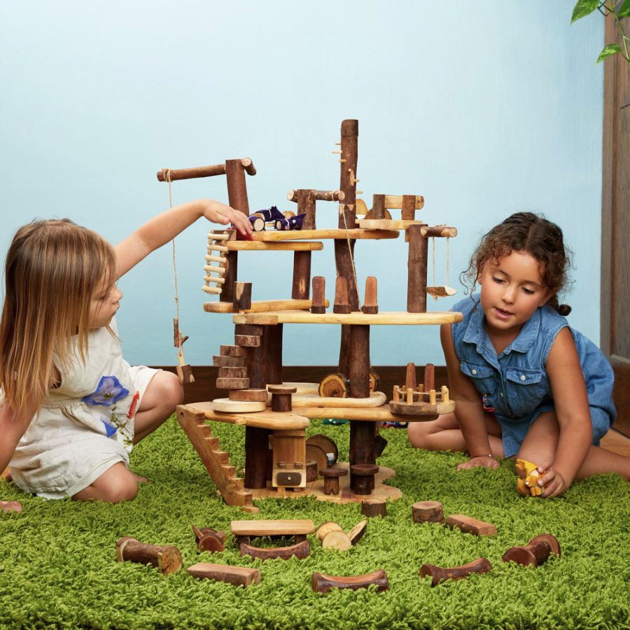 Kids playing with a Tree Blocks Build a Treehouse play structure made from natural logs and light wood boards, featuring platforms, stairs, hanging ropes, and small wooden toys.