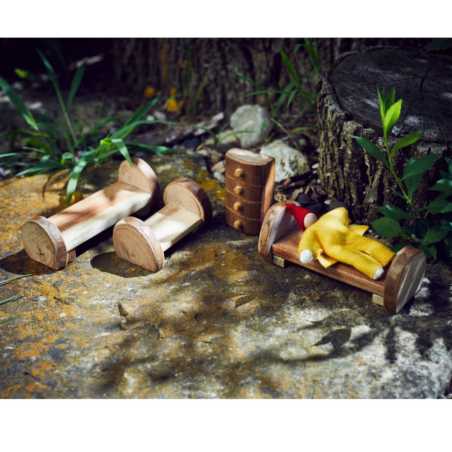 Tree Blocks bedroom furniture set including a small bed with yellow doll figure, two other beds, and a small dresser, displayed outdoors on a stone surface with plants and a tree stump in the background.