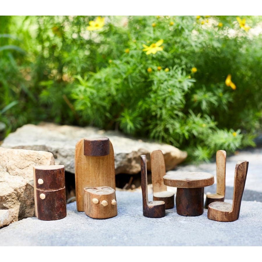 A set of small handcrafted wooden kitchen furniture pieces from Tree Blocks, including chairs, stools, and a table, arranged outdoors on a stone surface with a blurred green garden background.