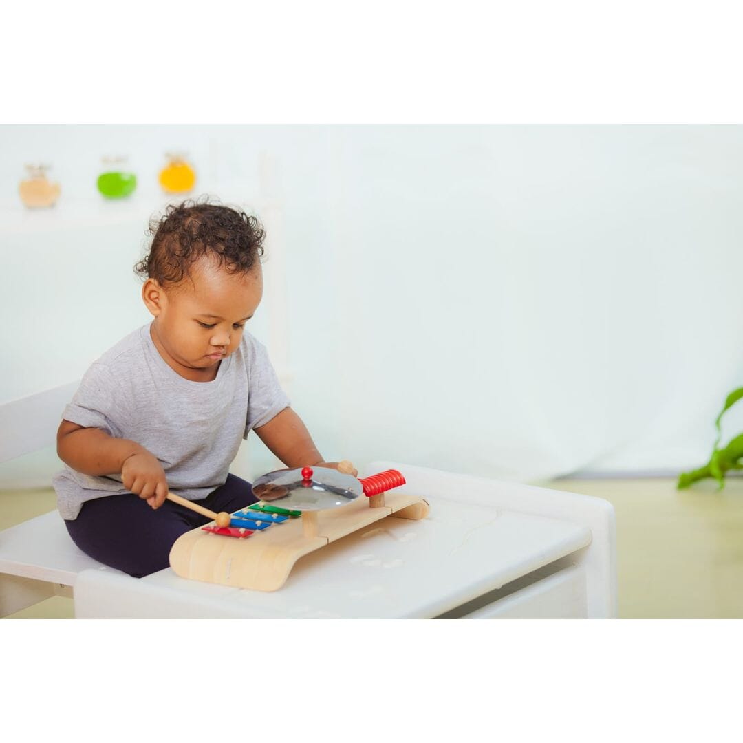 Small child plays a PlanToys Musical Set with rainbow xylophone, cymbals, and wooden guiro.