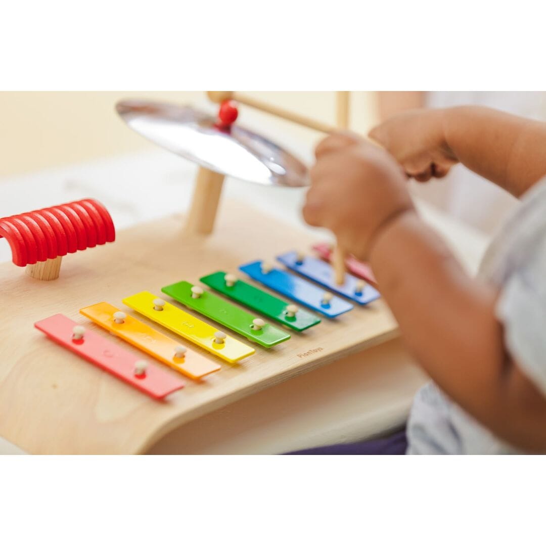 Close up small child plays a PlanToys Musical Set with rainbow xylophone, cymbals, and wooden guiro.